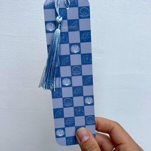 Könnte beinhalten: Ein blaues Lesezeichen mit einem karierten Muster und weißen Muscheln. Das Lesezeichen hat einen blauen Quasten.
