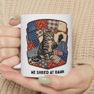 May include: White ceramic mug with a tabby cat illustration waving a paw, set against a patchwork quilt design in red, white, and blue. The text "WE SHRED AT DAWN" is printed below the cat.