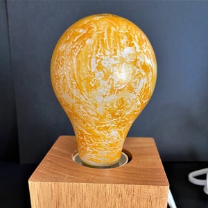 Puede incluir: Una bombilla amarilla con vetas blancas se asienta sobre una base de madera. La base es un bloque cuadrado de madera con un agujero en el centro para la bombilla.