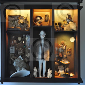 Puede incluir: Una figurita blanca de una persona vestida con un traje y una corbata se encuentra en una vitrina de varios compartimentos. La vitrina está llena de varios objetos, incluyendo un pequeño pájaro blanco, una botella de vidrio y un trozo de papel con escritura en él.