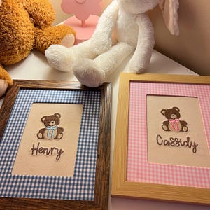 May include: Two framed embroidered pictures with teddy bear designs. One frame has a blue and white gingham border and the name "Henry". The other has a pink and white gingham border and the name "Cassidy".
