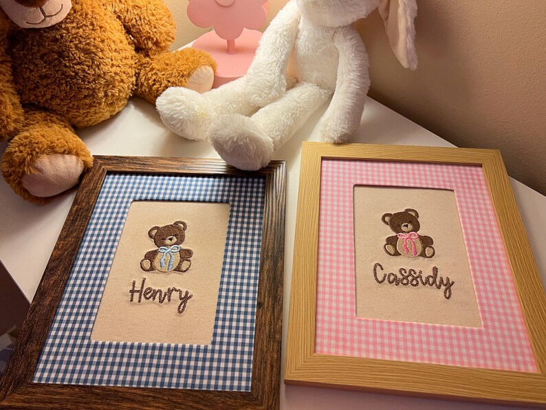 May include: Two framed embroidered pictures with teddy bear designs. One frame has a blue and white gingham border and the name "Henry". The other has a pink and white gingham border and the name "Cassidy".