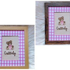 May include: Two framed pictures with a pink and white gingham pattern. Each frame holds a beige rectangle with a brown teddy bear embroidered on it, wearing a pink bow. The name "Cassidy" is embroidered below the bear. One frame is light wood, the other is dark wood.