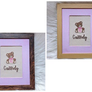 May include: Two framed embroidered pictures. Each features a brown teddy bear with a pink bow and the name "Cassidy" in script. The background is a light-colored fabric with a pink and white gingham pattern.