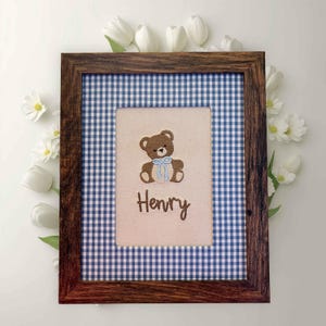 May include: A framed artwork featuring a brown teddy bear with a blue bow, embroidered on a beige background. The name "Henry" is stitched below. The frame is dark wood, set against a blue and white gingham pattern, and surrounded by white flowers.