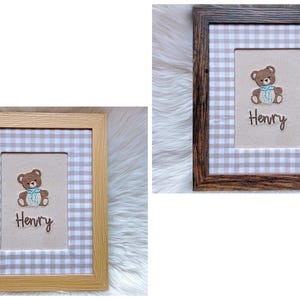 May include: Two framed baby nursery art pieces. One frame is light wood, the other is dark wood. Each features a teddy bear with a blue bow, the name "Henry", and a gingham pattern background. The frames are rectangular.