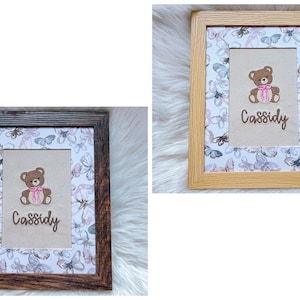 May include: Two framed pictures with a teddy bear design. The bear is brown with a pink bow and the name "Cassidy" is written below. The background features a butterfly pattern. One frame is light brown, the other is dark brown.