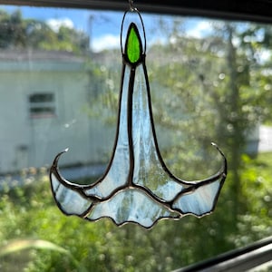 May include: A stained glass flower-shaped suncatcher with a green top and light blue petals. The glass is framed with a dark metal and hangs from a chain. The suncatcher is hanging in front of a window.