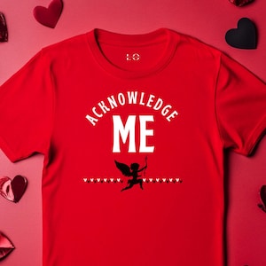 May include: A red t-shirt with the words "ACKNOWLEDGE ME" in white, above a black silhouette of Cupid aiming an arrow. Small red hearts are printed above and below the Cupid graphic. The shirt is surrounded by red and black heart decorations.