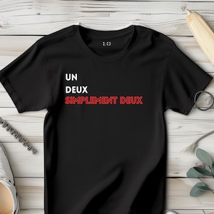 Könnte beinhalten: Schwarzes T-Shirt mit dem Text "UN DEUX SIMPLEMENT DEUX" in Weiß und Rot. Das Shirt wird auf einer hellen Holzoberfläche präsentiert, mit einem Paar weißer Turnschuhe, einem Bleistift und einer Brille im Hintergrund.
