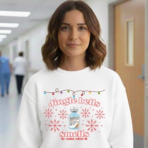 Könnte beinhalten: Ein weißes Sweatshirt mit einem festlichen Design mit dem Text "Jingle bells smells", einer Fläschchen-Grafik, Schneeflocken und Lichterketten. Das Sweatshirt wird von einer Person in einer Krankenhausumgebung getragen.
