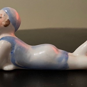 May include: A porcelain figurine of a woman in a blue swimsuit, lying on her stomach with her head turned to the side. The figurine is painted in shades of blue and white.