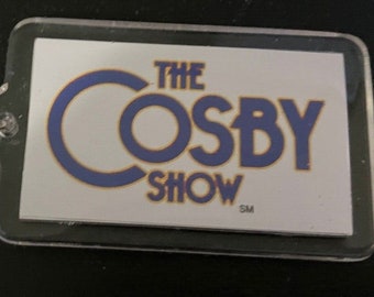 May include: A clear plastic keychain with a silver key ring. The keychain features a white rectangular tag with the text "THE COSBY SHOW" in blue and yellow lettering.