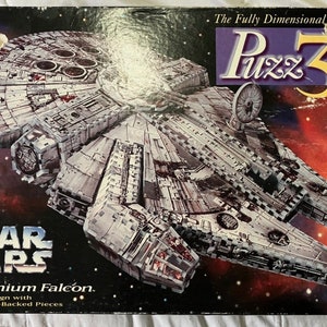 May include: A Star Wars Millennium Falcon 3D puzzle box. The box features a colorful image of the Millennium Falcon spaceship against a red and black space background. The text "The Fully Dimensional Puzzle" and "Puzz 3D" are printed on the top of the box. The text "Star Wars Millennium Falcon" is printed on the bottom of the box.
