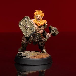 May include: A miniature figurine of a warrior with fiery orange hair and beard, holding a shield and a hammer. The warrior is clad in bronze armor and stands on a small, textured base. The background is a solid, deep red.
