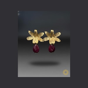 May include: A pair of gold-colored flower earrings with a teardrop-shaped, deep red gemstone pendant. The earrings have a detailed floral design and are set against a gradient gray background.