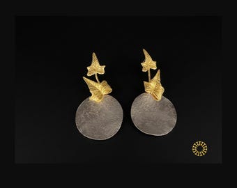 An outstanding bold fashionable earrings. They feature intricate, gold-toned ivy leaf motifs attached to textured, circular silver discs.