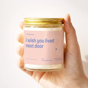 May include: A clear glass jar candle with a gold lid, held in a hand. The label is light pink with the text "i wish you lived next door" in blue. The candle is labeled "Tall Cup Of Candle" and "100% Soy Wax Candle".