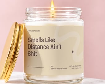 May include: A clear glass jar candle with a lit wick and a gold metal lid. The label features the text "Tall Cup Of Candle", "Smells Like Distance Ain't Shit", "V132", "Scent 03, White Tea + Jasmine", "100% Soy Wax Candle", and "Made in California".