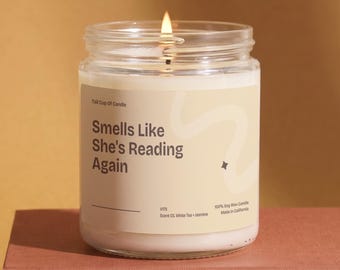 Op de afbeelding: Een kaars in een doorzichtige glazen pot met witte was en een brandende lont. Het crèmekleurige etiket bevat de donkerbruine tekst: Tall Cup Of Candle, Smells Like She's Reading Again, V175, Scent 03, White Tea + Jasmine, 100% Soy Wax Candle.