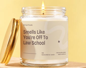 Peut inclure: Une bougie en pot de verre transparent avec une mèche allumée et un couvercle en métal doré. L'étiquette porte le texte "Smells Like You're Off To Law School", "Tall Cup Of Candle", "V222", "Scent 03. White Tea + Jasmine" et "100% Soy Wax Candle".