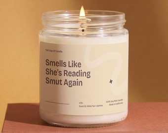 Peut inclure: Une bougie en pot de verre transparent avec de la cire blanche et une mèche allumée. L'étiquette porte le texte "Tall Cup Of Candle," "Smells Like She's Reading Smut Again," "V176," "Scent 03, White Tea + Jasmine," "100% Soy Wax Candle," et "Made in California."