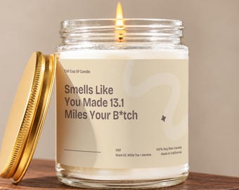 May include: A clear glass jar candle with a lit wick and a gold-colored metal lid. The label features the text "Smells Like You Made 13.1 Miles Your B*tch" along with "Tall Cup Of Candle", "V197", "Scent 03. White Tea + Jasmine", and "100% Soy Wax Candle".