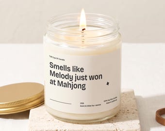 Puede incluir: Una vela en frasco de vidrio transparente con una mecha encendida y una etiqueta blanca con el texto en negro: Tall Cup Of Candle, Smells like Melody just won at Mahjong, V1126, Scent 03, White Tea + Jasmine, 100% Soy Wax Candle, Made in California.