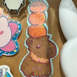 May include: A sticker featuring a brown capybara with a stack of orange pumpkins on its head. The sticker has a light blue border and holographic accents. Other stickers are visible in the background.