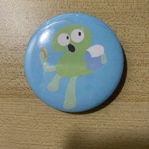May include: A blue and green button with a cartoon character on it. The character is a green number two with a surprised expression. The character is holding a pencil in its right hand and a blue bucket in its left hand.