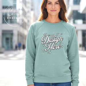以下が含まれることがあります： 白文字で「Your Design Here」と書かれた、薄緑色のラグランスリーブのスウェットシャツ。 スウェットシャツは無地で、クルーネックです。 モデルはぼやけた背景の前に立っています。