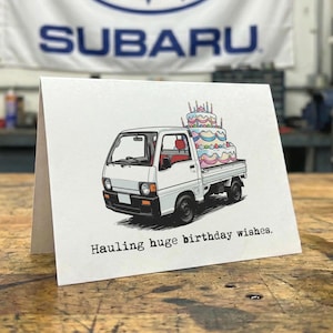 May include: Greeting card featuring a white truck carrying a colorful, multi-tiered birthday cake. The card reads "Hauling huge birthday wishes." A Subaru banner is visible in the background.