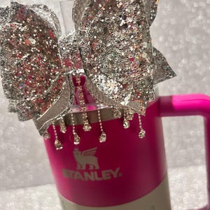 May include: A pink Stanley travel mug with a silver glitter bow and rhinestone accents. The mug has the Stanley logo and the text "Quencher H2O Tumbler" on it.