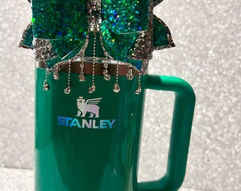 Op de afbeelding: Een groene Stanley-reistumbler met een zilveren logo en een groene glitter strik met zilveren versiering en een ketting van zilveren bedels.