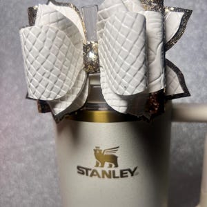 Peut inclure: Un nœud blanc matelassé avec des accents de paillettes dorées est posé sur un gobelet Stanley crème. Le nœud est fait d'un matériau texturé et est attaché à une paille transparente. Le logo Stanley est visible sur le gobelet.