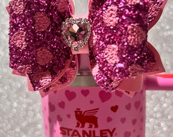 May include: Pink Stanley travel mug with a pink glitter heart-shaped bow with a rhinestone heart in the center. The mug has a white logo that says "STANLEY" and is covered in pink hearts.