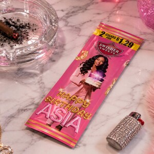 K&ouml;nnte beinhalten: Eine rosa Swisher Sweets Zigarrenpackung mit einer Frau, die einen Kuchen h&auml;lt, dem Text "Happy Birthday Asia" und einem Preisschild. Eine brennende Zigarre liegt in einem durchsichtigen Aschenbecher. Ein mit Strass besetztes Feuerzeug und Schmuck sind ebenfalls sichtbar.