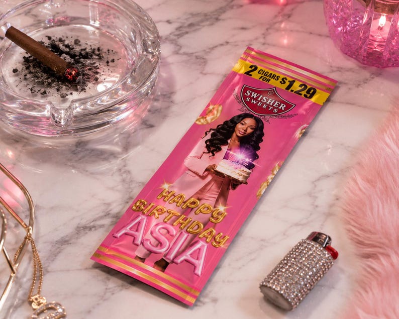 K&ouml;nnte beinhalten: Eine rosa Swisher Sweets Zigarrenpackung mit einer Frau, die einen Kuchen h&auml;lt, dem Text "Happy Birthday Asia" und einem Preisschild. Eine brennende Zigarre liegt in einem durchsichtigen Aschenbecher. Ein mit Strass besetztes Feuerzeug und Schmuck sind ebenfalls sichtbar.