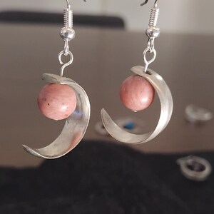 May include: A pair of silver earrings with a hook closure. Each earring features a pink, spherical bead cradled by a curved silver element. The earrings are displayed against a blurred background.