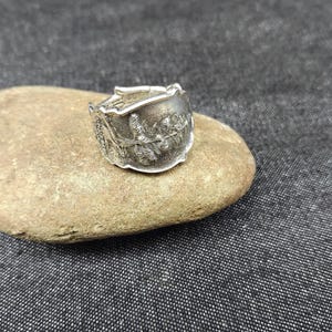 May include: A silver ring with a floral design. The ring appears to be made from a spoon, with the handle forming the band and the bowl of the spoon creating the face of the ring. The ring is resting on a smooth, light brown stone.