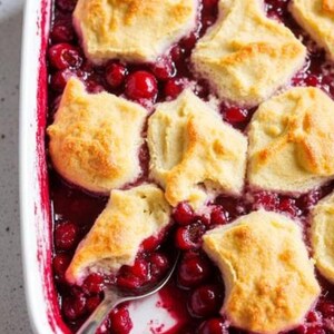 May include: A close-up shot of a baked cherry cobbler in a white rectangular dish. The cobbler features golden-brown biscuits atop a vibrant red cherry filling. A silver spoon rests in the dish, partially submerged in the cherry sauce.