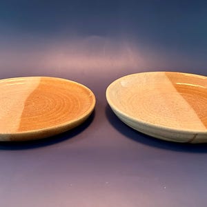Pode incluir: Dois pratos redondos de cerâmica com um design de dois tons, apresentando uma secção de cor creme clara e uma secção de cor bege quente. Os pratos têm uma textura lisa e ligeiramente salpicada, com uma base larga e pouco profunda.