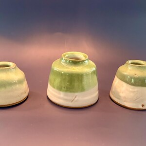 May include: Three ceramic vessels with a two-tone glaze featuring a light green upper section and a white lower section. Each piece has a wide base, a tapered body, and a small circular opening at the top, displaying a smooth, glossy finish.