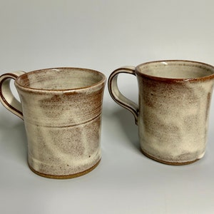 May include: Two ceramic mugs with brown rims and a white glaze. The mugs have a speckled brown and white pattern.
