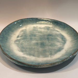May include: A round ceramic plate featuring a reactive glaze with a blend of deep teal and white colors. The center of the plate has a mottled white appearance, transitioning into a darker teal rim with a visible brown edge along the circumference.