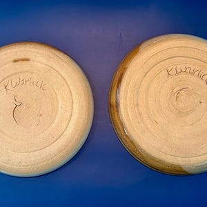 Może przedstawiać: Dwa okrągłe talerze ceramiczne widziane od spodu, z jasnobeżową, nieszkliwioną podstawą z gliny o koncentrycznych, kolistych teksturach. Każdy talerz ma ręcznie wyryty podpis K.Warlick na środku dolnej powierzchni.
