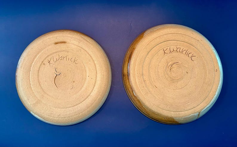 Może przedstawiać: Dwa okrągłe talerze ceramiczne widziane od spodu, z jasnobeżową, nieszkliwioną podstawą z gliny o koncentrycznych, kolistych teksturach. Każdy talerz ma ręcznie wyryty podpis K.Warlick na środku dolnej powierzchni.