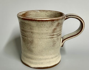 Może przedstawiać: Biały kubek ceramiczny z brązową krawędzią i uchwytem. Kubek ma rustykalny, ręcznie robiony wygląd.