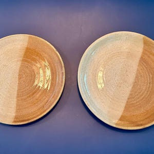Może przedstawiać: Dwa okrągłe talerze ceramiczne o dwukolorowym wzorze. Każdy talerz ma nakrapianą, kremowobiałą glazurę z jednej strony i gładką, jasnobrązową glazurę z drugiej, co tworzy wyraźny podział na powierzchni tego naczynia stołowego.