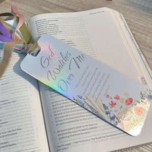 May include: A holographic bookmark with the text "God Watches Over Me" and floral accents. The bookmark has colourful ribbons attached and rests on an open book. A small succulent plant is in the background.